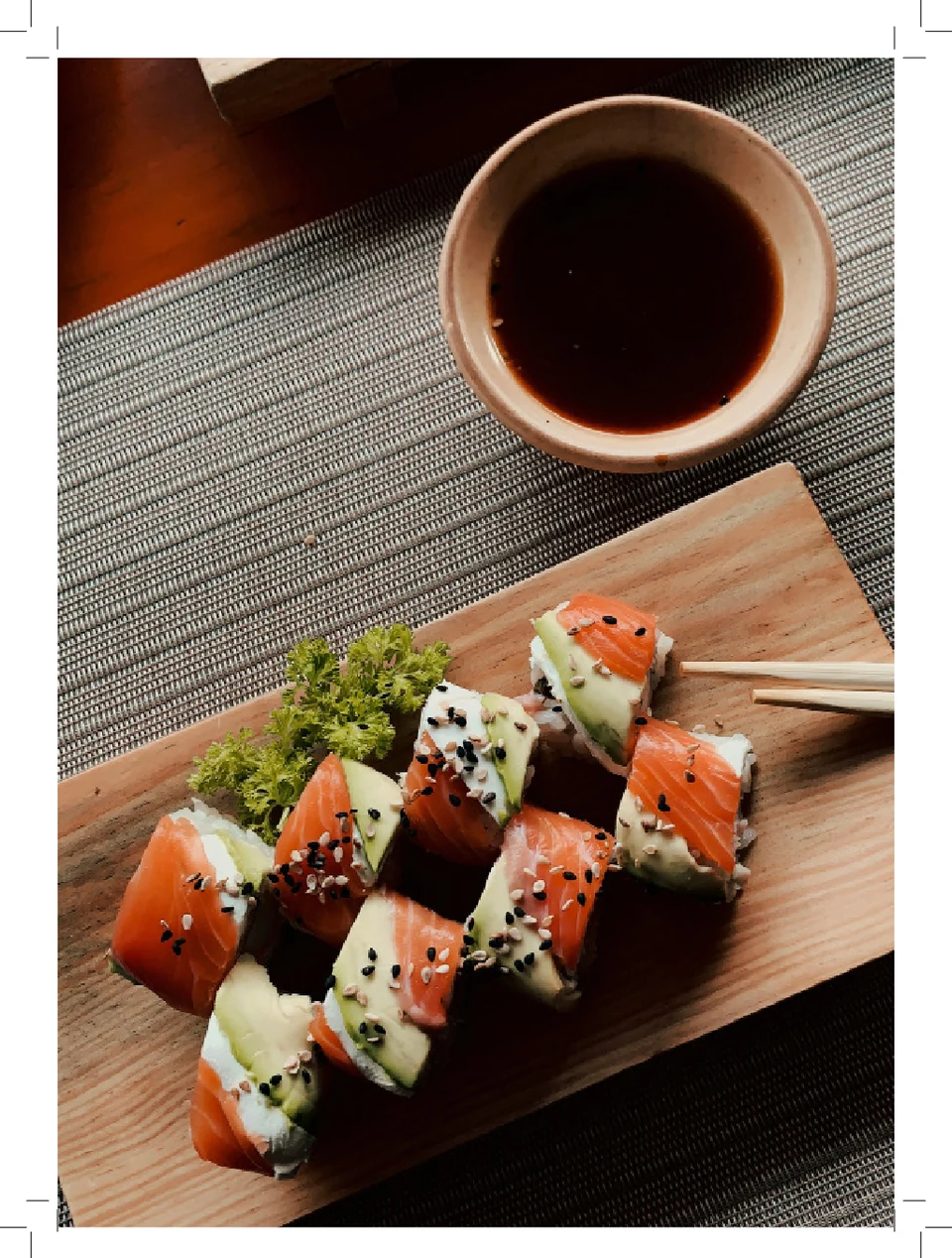 Salmon and avocado sushi on a wooden board beside a cup, photographed from above within a white print border.